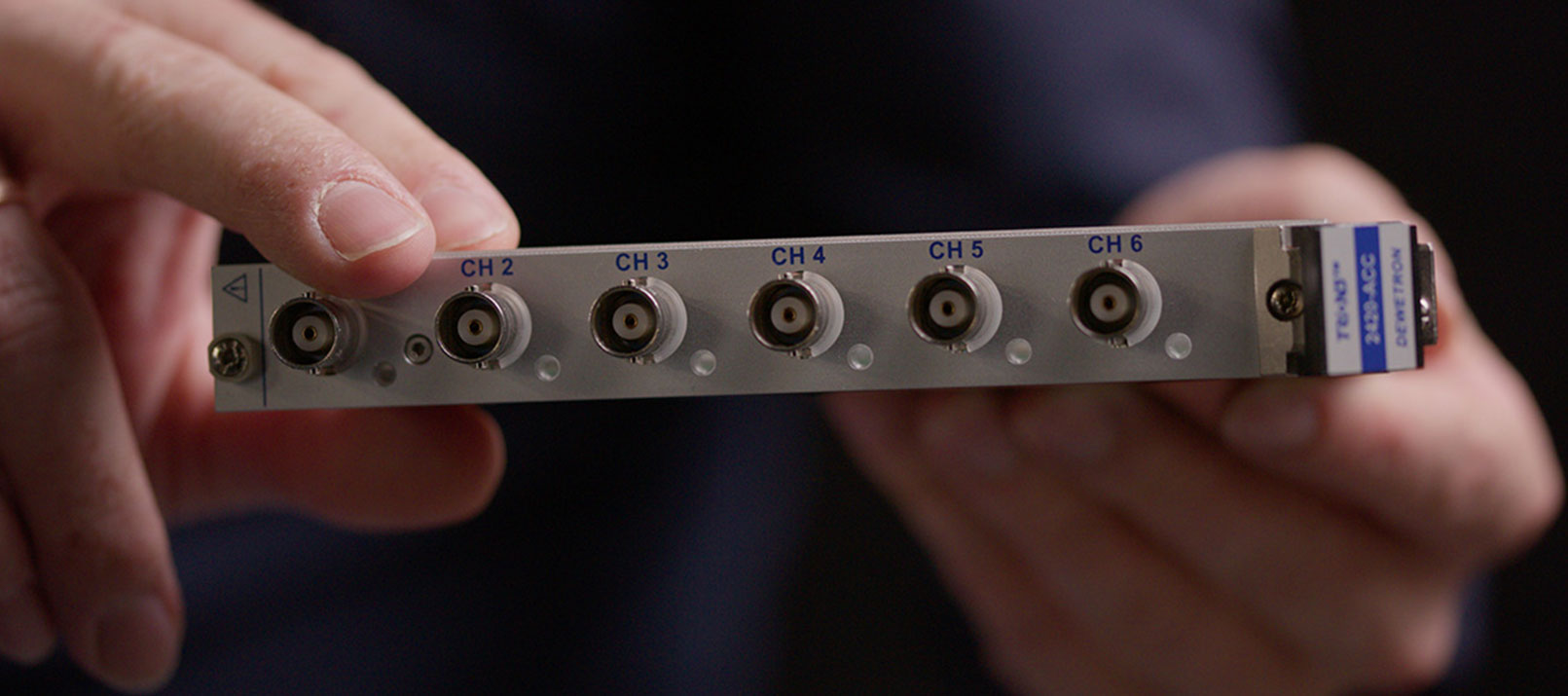 A person holds a trion3-24xx-acc-lv metal panel with six labeled coaxial connectors, marked CH 1 to CH 6, and a blue and white label on the right side. The panel is shown against a dark, blurred background.