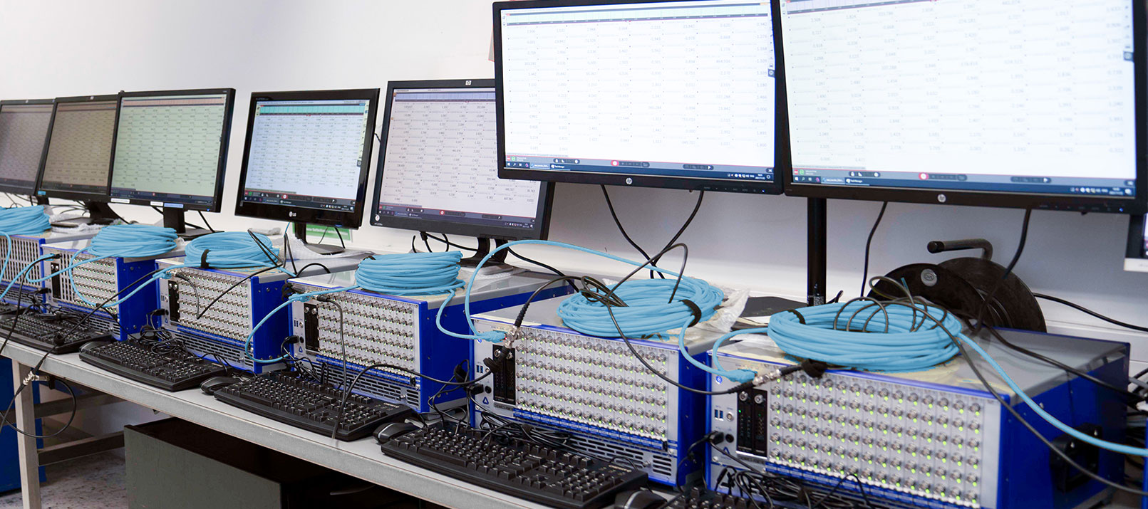 A row of computers with multiple screens, keyboards, and large blue ethernet cables on a workbench in a lab or data center setting supports distributed measurements for OXYGEN-NET. The monitors display spreadsheets or data tables.