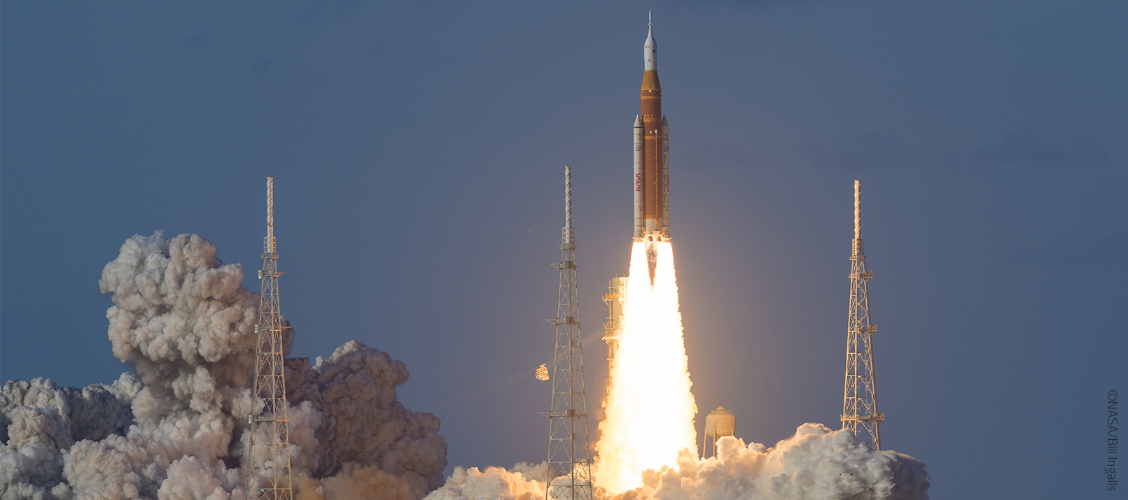 Eine große Rakete der NASA-Mission Artemis startet in den Himmel und lässt helle Flammen und Rauch aufsteigen. Vier Starttürme umgeben die Rakete, während im Hintergrund ein klarer Himmel und ein Hauch von Wolken zu sehen ist.