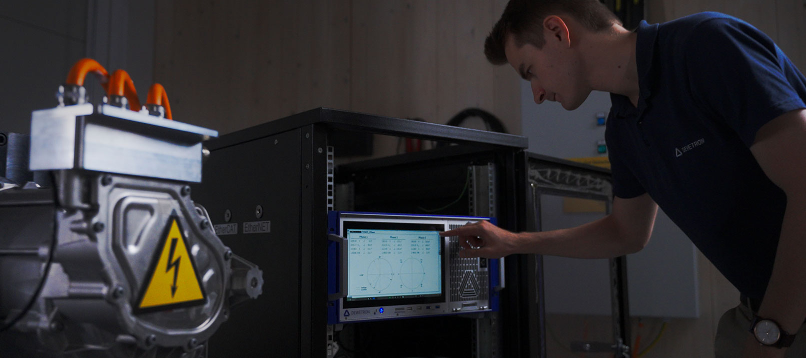 A person operates a touchscreen panel on an electrical testing device in a lab, with machinery featuring a high voltage warning symbol visible in the foreground.