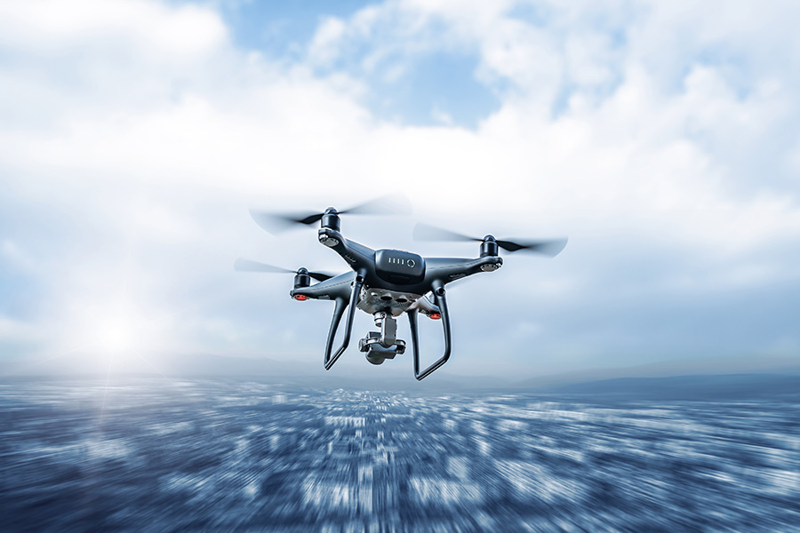 A black quadcopter drone with a camera, utilized for Aerospace and Aviation Testing, flies above a blurred cityscape under a cloudy sky, with the sun shining in the background.