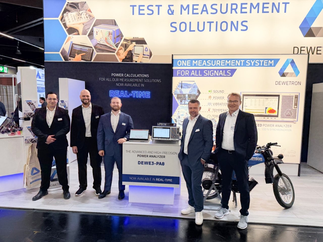 Vier Männer in Anzügen stehen an einem DEWETRON-Messestand, an dem ein Leistungsanalysator vom Typ DEWE3-PA8 und ein Motorrad ausgestellt sind.